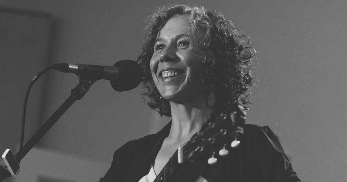 In black and white, Ann is smiling while playing a guitar and standing in front of a stage microphone.