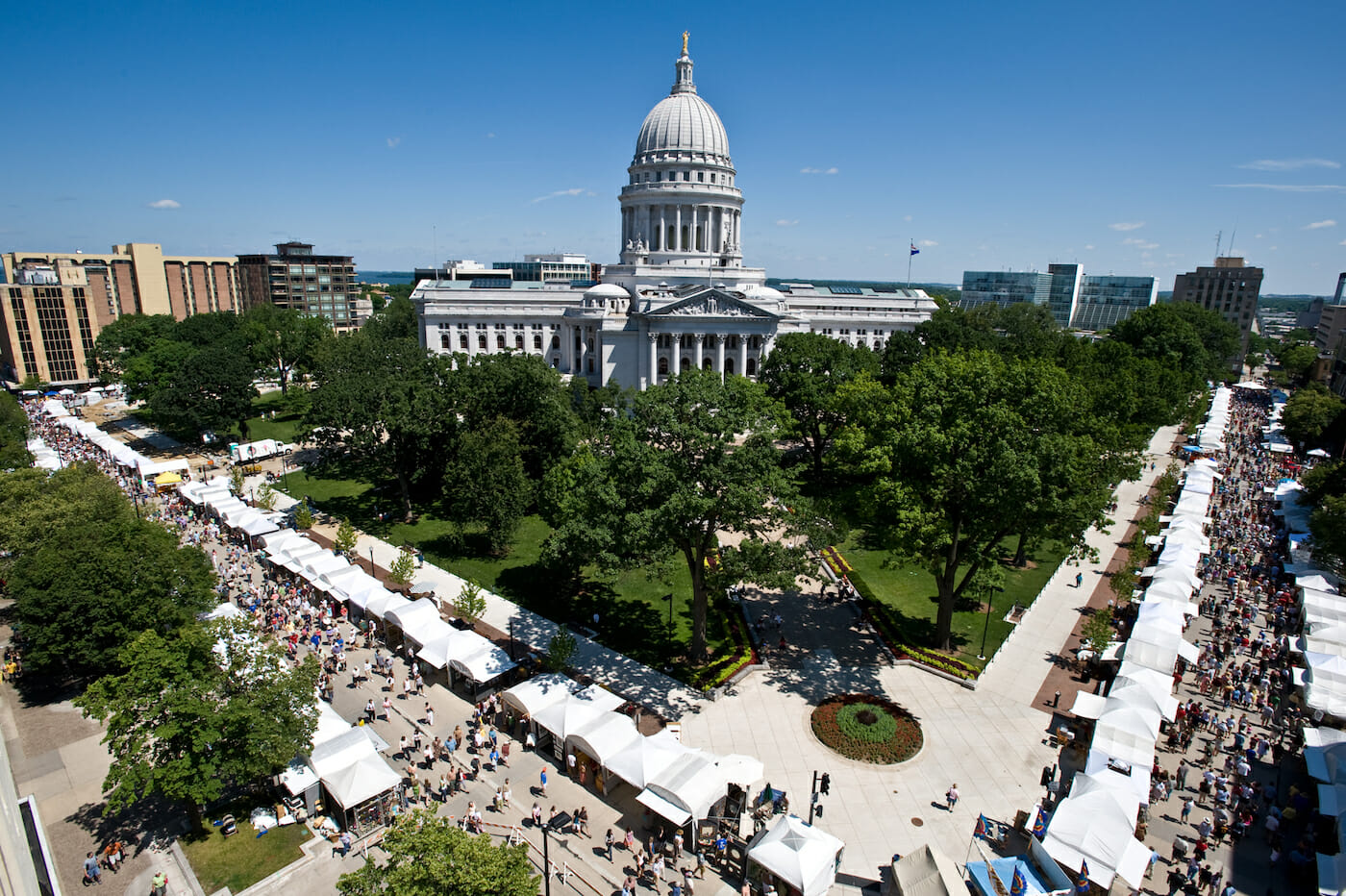 An aerial view of the Wisconsin Capitol building surrounded by art fair booths and crowds of people. The sky is sunny and blue.