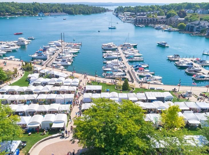 A bay filled with boats is seen from above. In the foreground, white art fair booths are lined up in neat rows on a lawn.